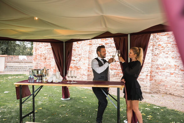 Unter dem eleganten Faltpavillon mit Himmel stehen 2 Personen mit einem Glas Prosecco in der Hand. 