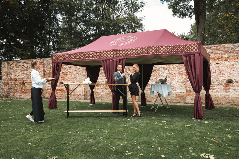 Das bordeaux-farbene Cateringzelt steht in einer Wiese. Davor steht ein Pärchen und hält ein Glas Prosecco in der Hand. Die Servicefachkraft bringt Häppchen auf einem Tablet.