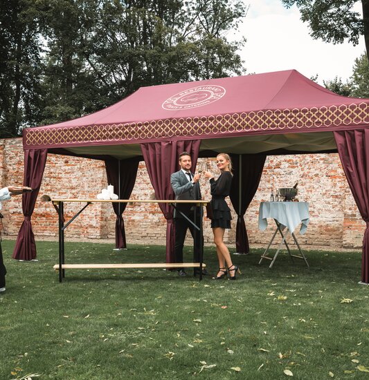 Das bordeaux-farbene Cateringzelt steht in einer Wiese. Davor steht ein Pärchen und hält ein Glas Prosecco in der Hand. Die Servicefachkraft bringt Häppchen auf einem Tablet.