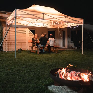 Das 4,5x3 m Gartenzelt steht im Dunkeln im Garten. Darunter sitzen die 4 Gäste. Vor dem Zelt ist noch eine Feuerstelle.