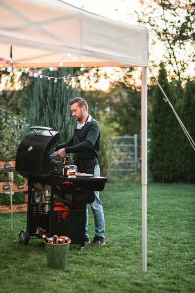 Der Mann steht mit seinem Grill unter einem ecru-farbenen Faltpavillon im Garten. Der Faltpavillon ist dekoriert mit Lichterketten und gesichert mit Spanngurten.